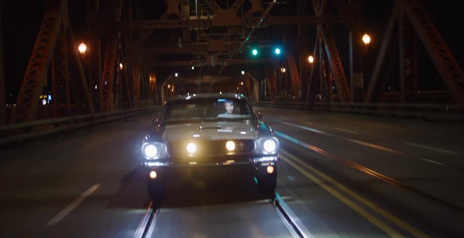 A woman drives a classic muscle car across an illuminated bridge at night, with the camera tilting down from the steel girders