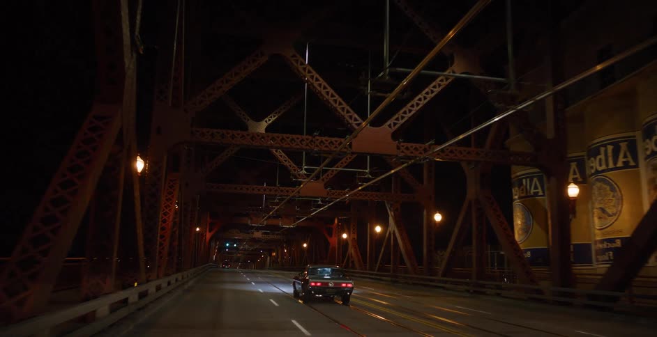 A vintage muscle car drives across a large industrial steel bridge at night
