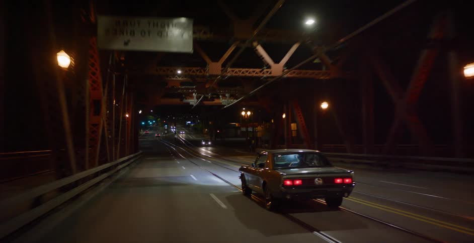 Following a classic muscle car as it drives over an illuminated industrial bridge and onto city streets at night