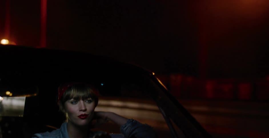 A stylish woman with red lipstick and a bandana sits pensively in her car on a dark street at night