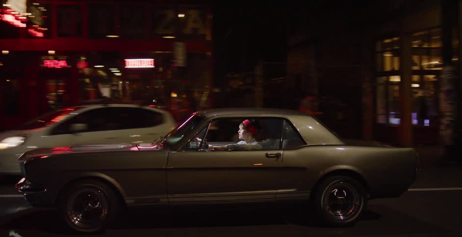 A stylish woman with a red bandana drives her classic American muscle car through the city at night