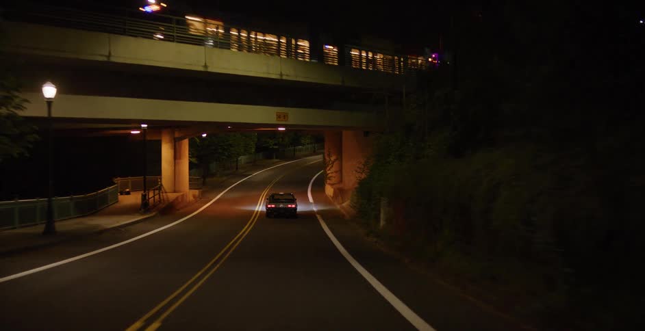 A vintage car travels along a dark, winding road under an overpass at night