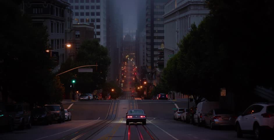 A vintage car slowly drives down a steep, foggy San Francisco street at night with city lights illuminating the way