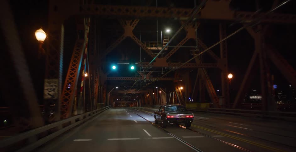 A classic muscle car drives over an illuminated steel truss bridge in a city at night
