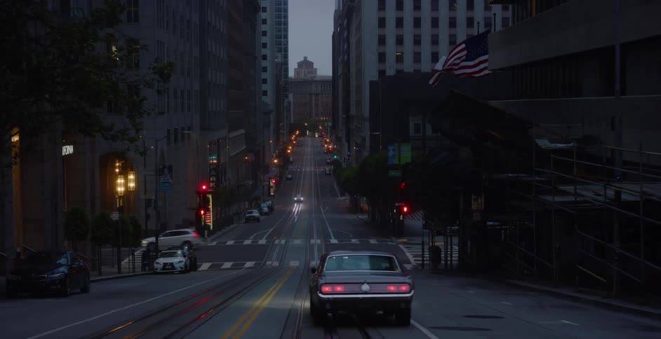 A vintage black muscle car drives down a steep, quiet city street in San Francisco at twilight