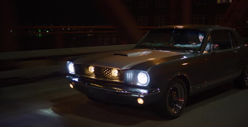 A stylish woman drives a classic grey Ford Mustang in slow motion across a city bridge at night