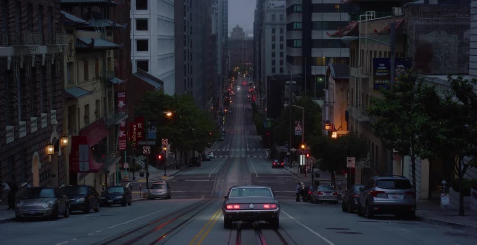 A classic American muscle car drives away down a steep, empty San Francisco street at twilight