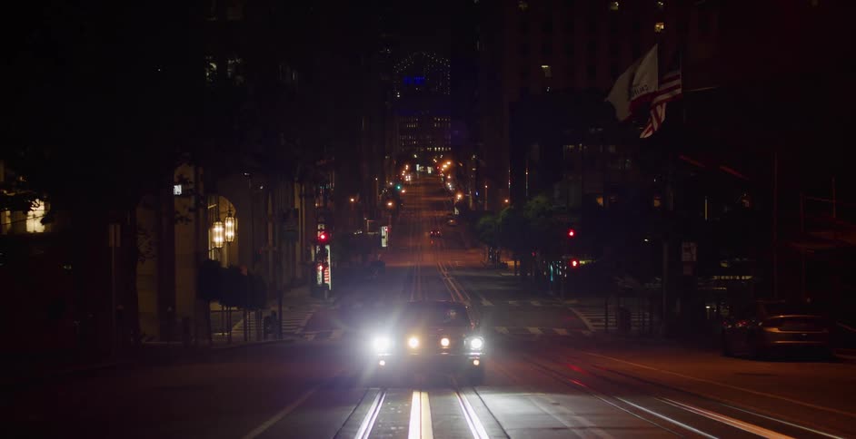 A car with bright headlights sits parked on a steep, empty San Francisco street with trolley tracks at night as the camera slowly zooms out