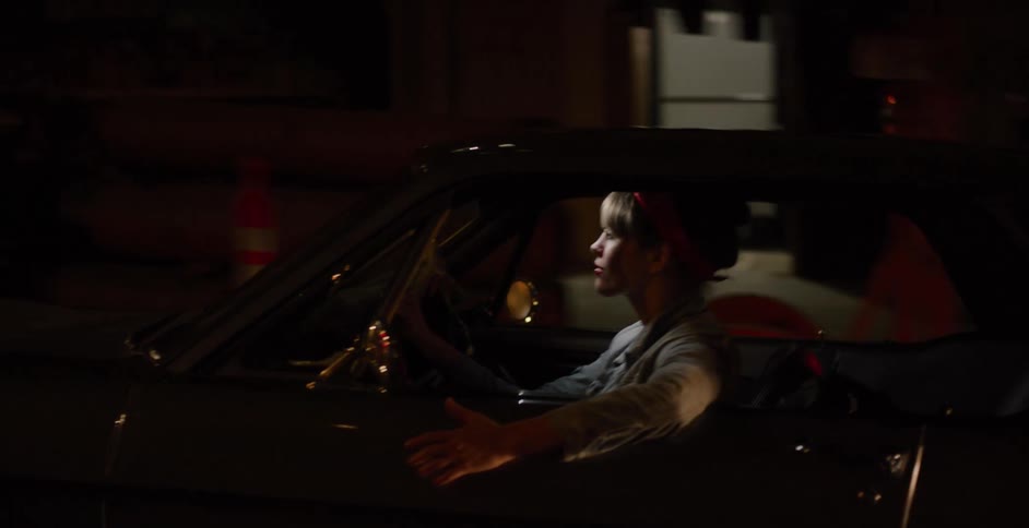 A stylish woman with a red bandana drives a classic car through the city at night, her arm resting on the open window