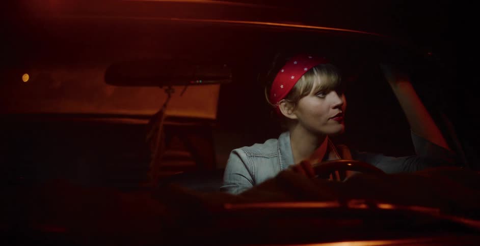 A stylish woman wearing a red bandana drives her car through the city at night as lights reflect off the windshield