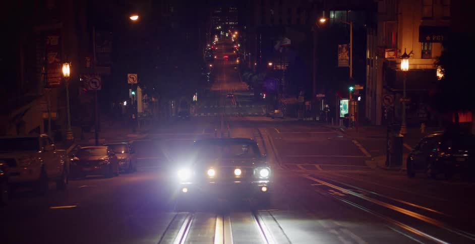 Slow zoom out reveals a vintage car with its headlights on, parked on the trolley tracks of a steep city street at night
