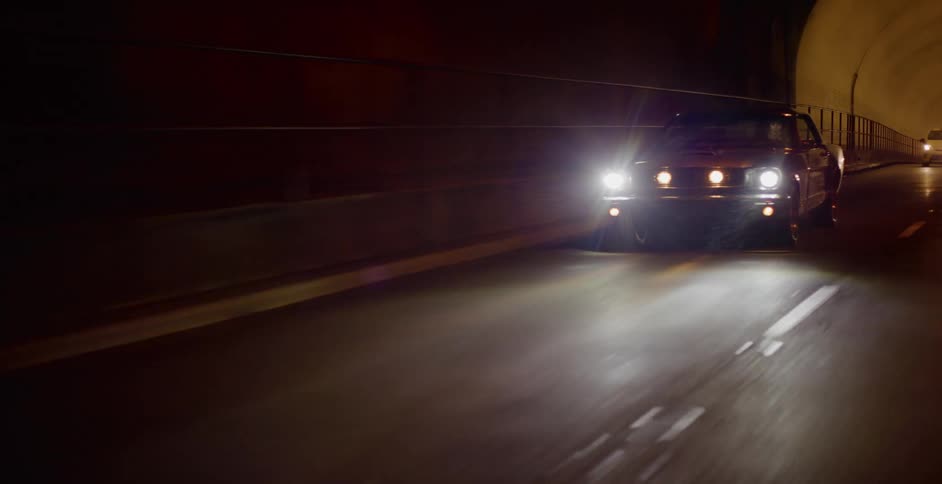 Front view of a classic silver American muscle car driving through a dark tunnel at night with its headlights on