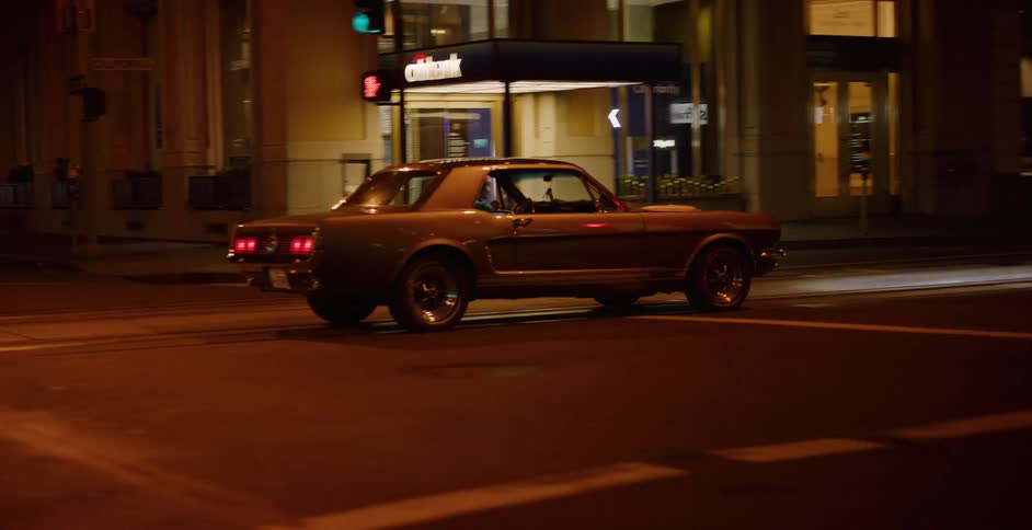 A vintage Ford Mustang drives through downtown city streets at night