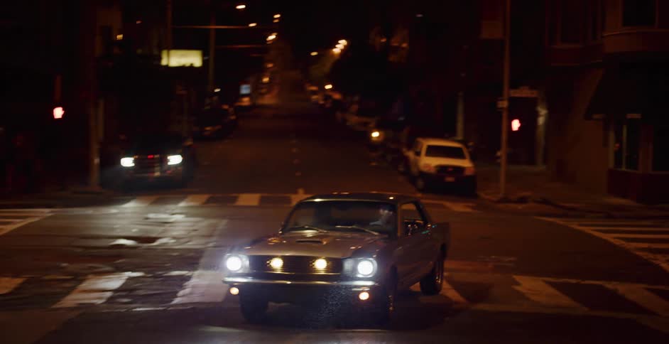 A classic silver Mustang with bright headlights cruises through a dark city intersection at night