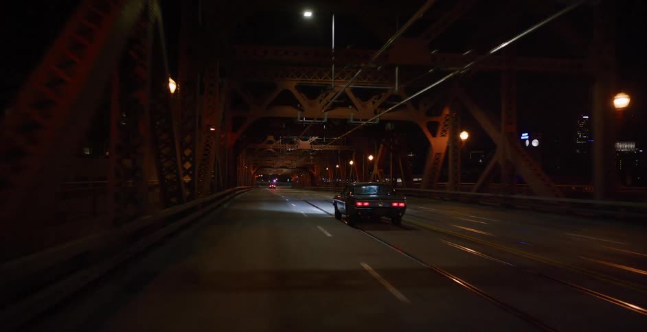 Following a vintage car while driving across a steel truss bridge illuminated by city lights at night