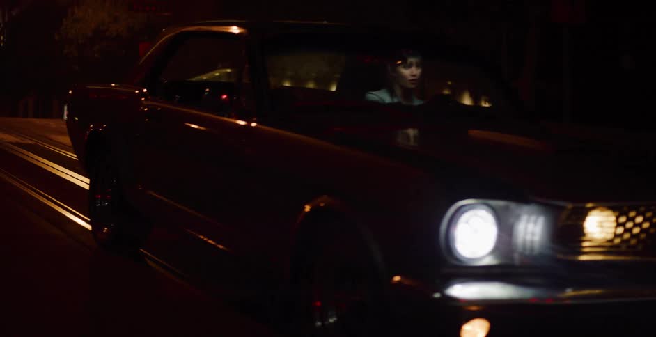 A classic black muscle car with its headlights on drives down a city street at night, revealing a woman behind the wheel