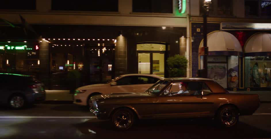 Side view of a person driving a classic Ford Mustang down a city street at night, passing by neon-lit storefronts and bars