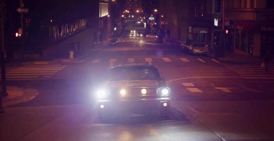 A classic muscle car with its headlights glaring drives down an empty city street late at night