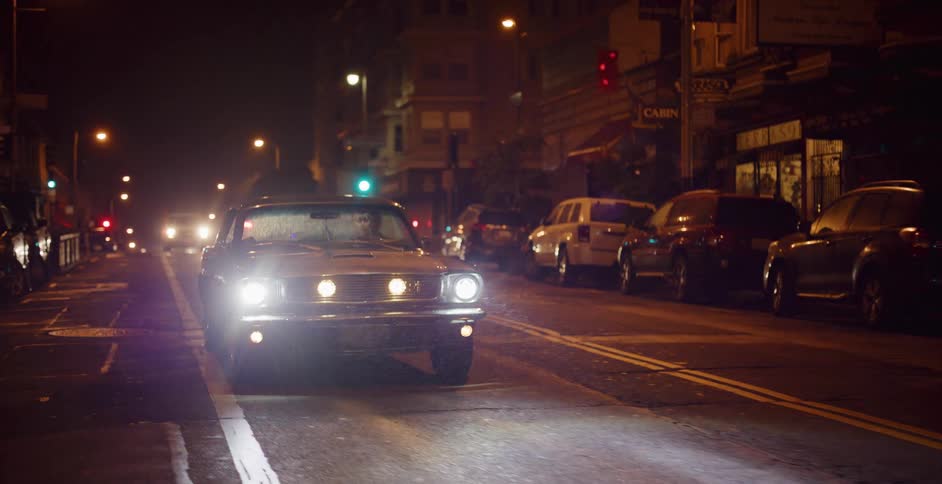 Front view of a classic muscle car with bright headlights driving slowly down a city street at night