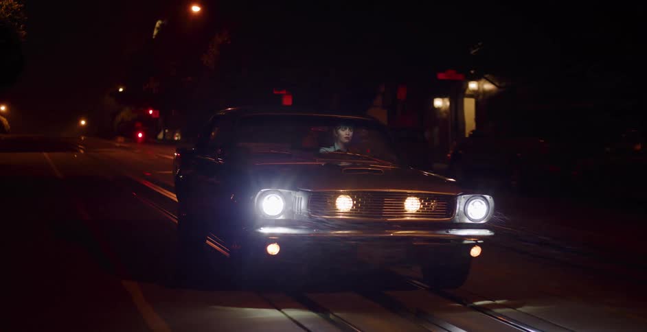 A woman with dark hair drives a classic muscle car with bright headlights down a wet city street at night