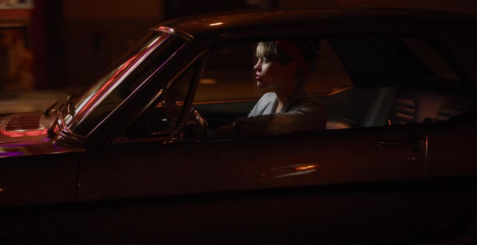 A stylish woman with a red bandana drives a classic muscle car through the city at night