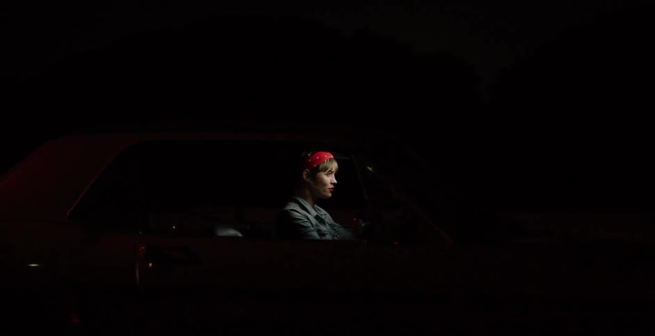 A stylish woman in a red bandana drives her classic orange muscle car down a dark street at night