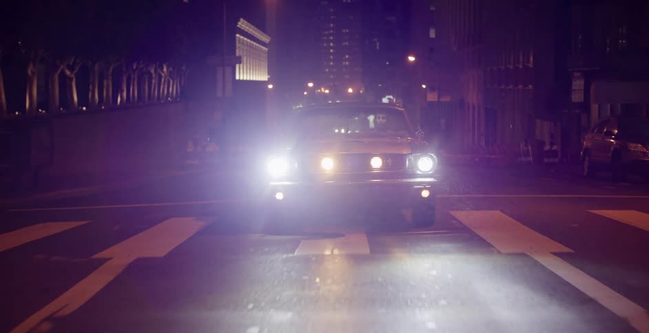 A classic muscle car slowly approaches on a wet city street at night with its bright headlights on