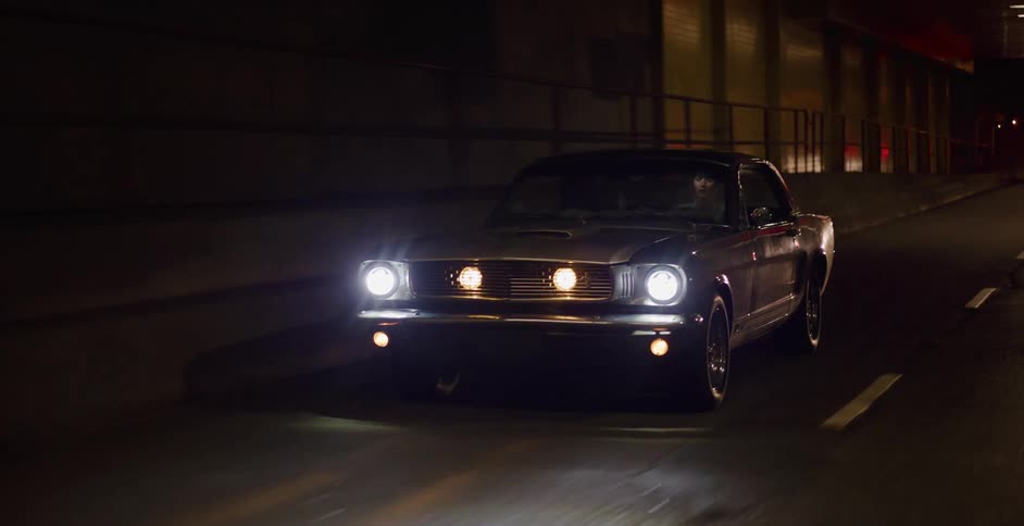 A stylish woman drives a classic silver Ford Mustang in slow motion through a dimly lit city tunnel at night