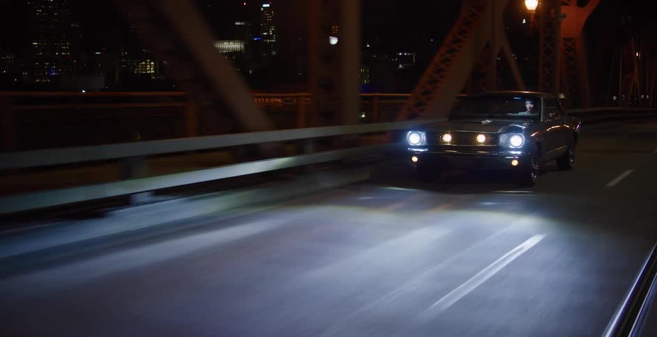 A woman drives a vintage muscle car across an illuminated city bridge at night, with the skyline in the background