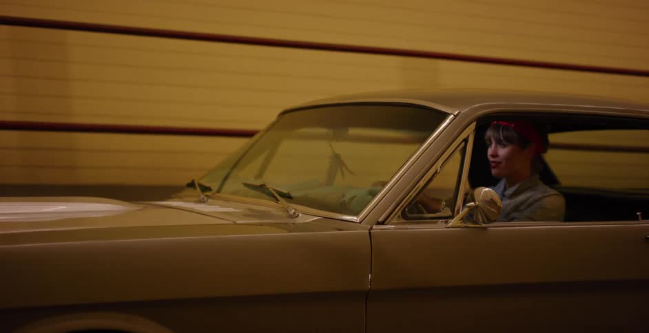 A stylish young woman with a red bandana drives a classic silver muscle car through a brightly lit tunnel at night