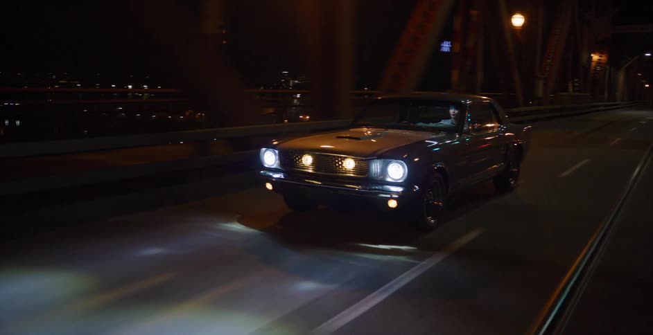 A classic grey Ford Mustang drives across an industrial metal bridge in the city at night
