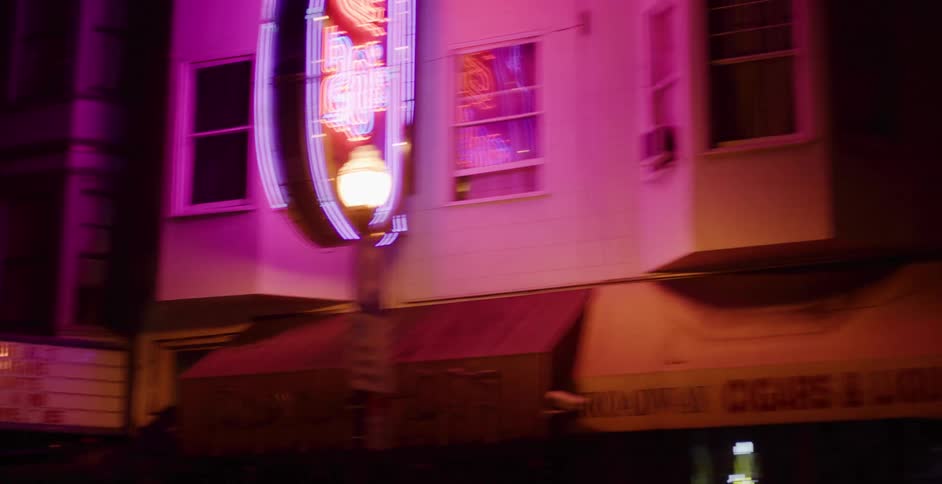 Vibrant neon signs of bars and clubs cast a colorful glow on a city street during a slow tracking shot at night