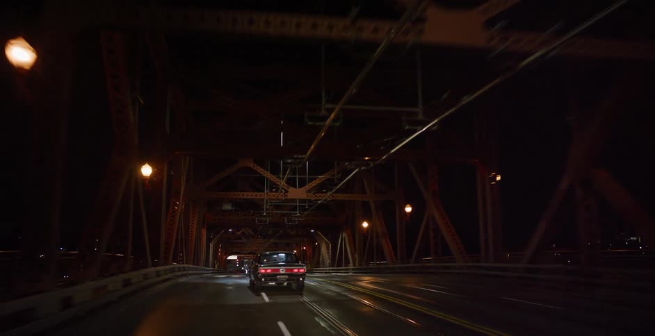 Following a vintage car as it drives over an industrial steel bridge in the city late at night