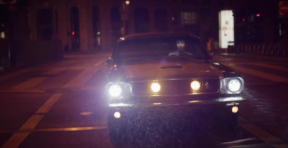 A woman drives a classic muscle car with bright headlights through misty city streets at night