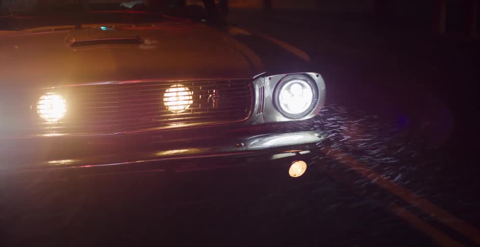 Close-up of a vintage muscle car with its headlights on during a rainy night