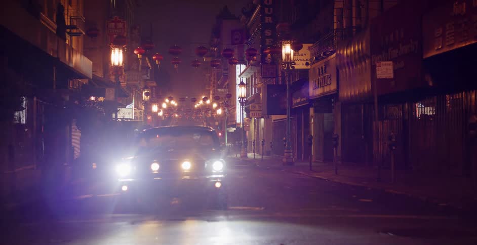 A vintage American muscle car with bright headlights sits on an empty, wet city street in Chinatown at night