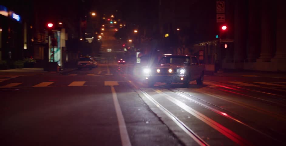 Camera pulls back from a classic Ford Mustang parked on a dark, empty city street at night with its headlights on