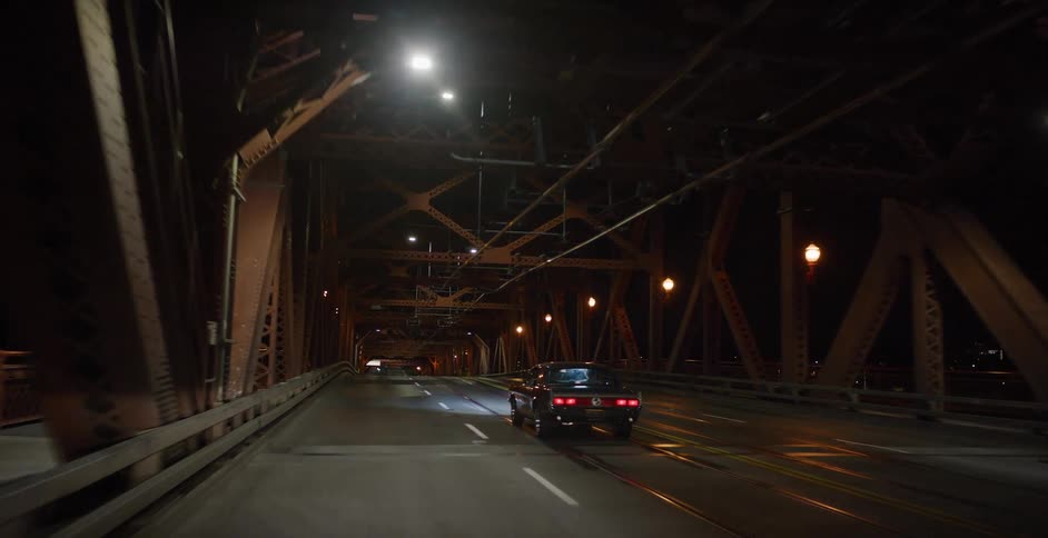 A vintage black car is followed across a metal truss bridge in the city late at night