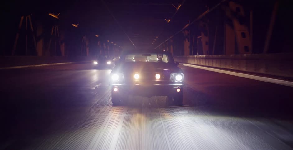 A man drives a classic muscle car with bright headlights across a bridge in the darkness