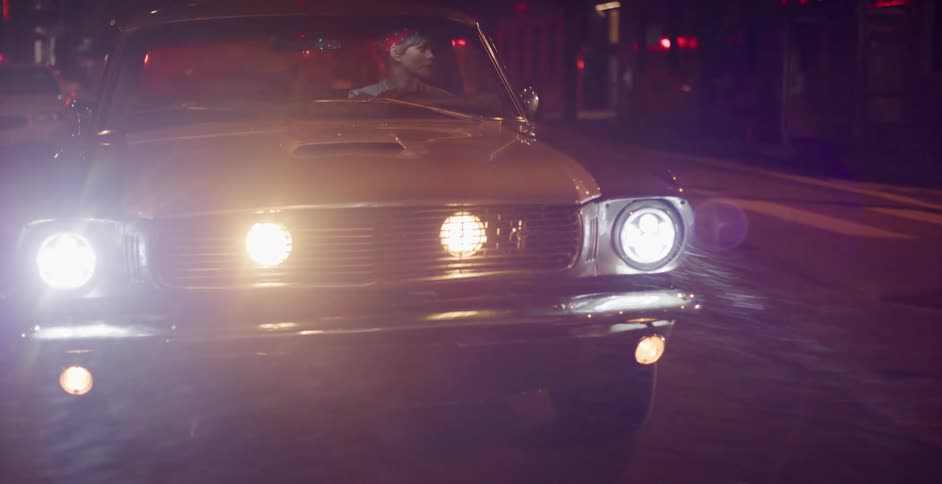 A stylish woman with sugar skull makeup drives her classic muscle car through the rainy city streets at night