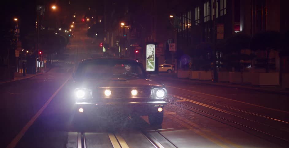 A classic muscle car with bright headlights drives slowly towards the camera on an empty city street at night