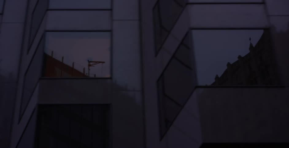 A slow pan reveals reflections of a city skyline and construction crane in the windows of a modern building at dusk