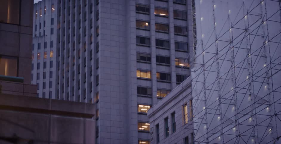 A slow dolly shot reveals the lit windows of city skyscrapers at dusk, with one building under construction wrapped in scaffolding