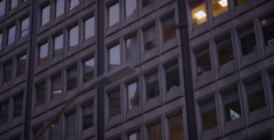 A slow pan across the monolithic facade of a city office building at dusk, with a few lit windows showing signs of life