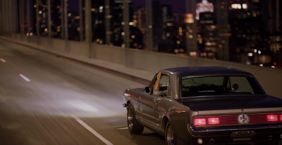 A classic Ford Mustang cruises over a city bridge at night, with the illuminated skyline in the background