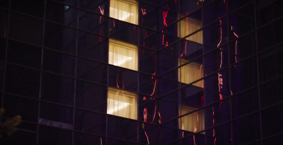 Red lights reflect on the glass facade of a modern skyscraper at night, with several windows brightly lit