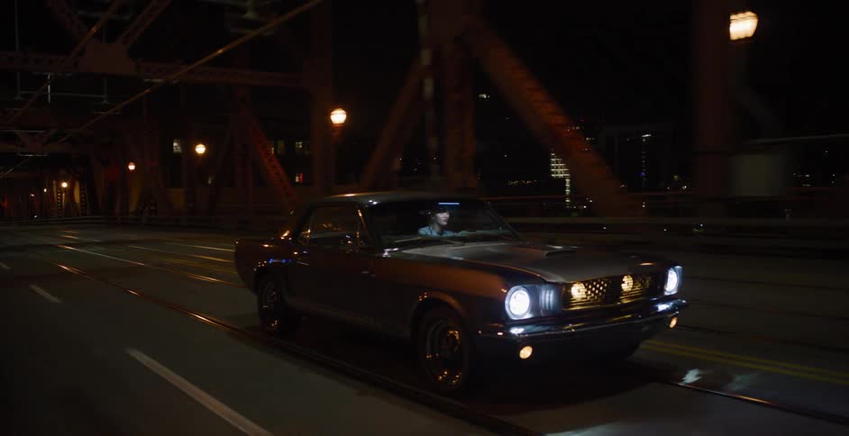 A man drives a classic muscle car across an illuminated steel truss bridge at night in the city