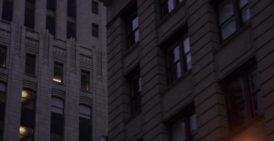A slow tilt down city skyscrapers at dusk reveals a street lamp turning on