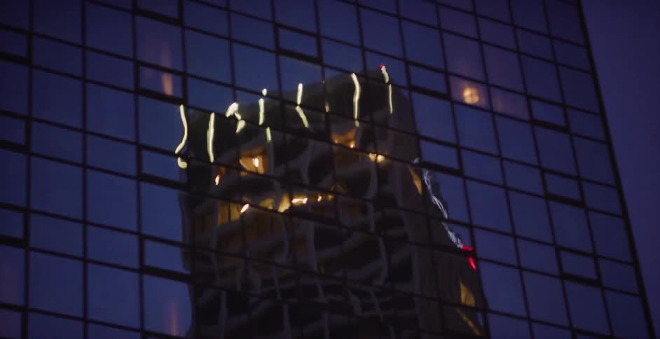 The distorted reflection of a modern skyscraper illuminates the windows of a glass building at night