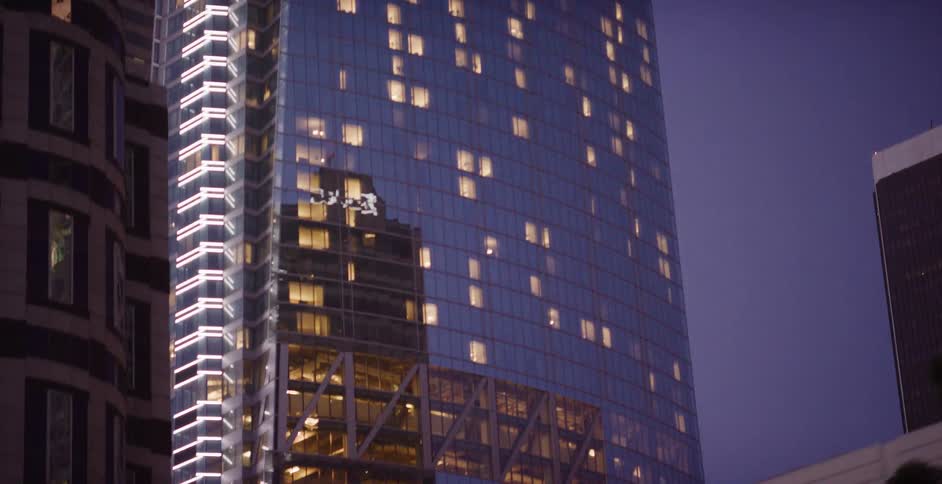 A slow downward pan of a modern glass skyscraper with glowing windows reflecting the city against a deep blue twilight sky